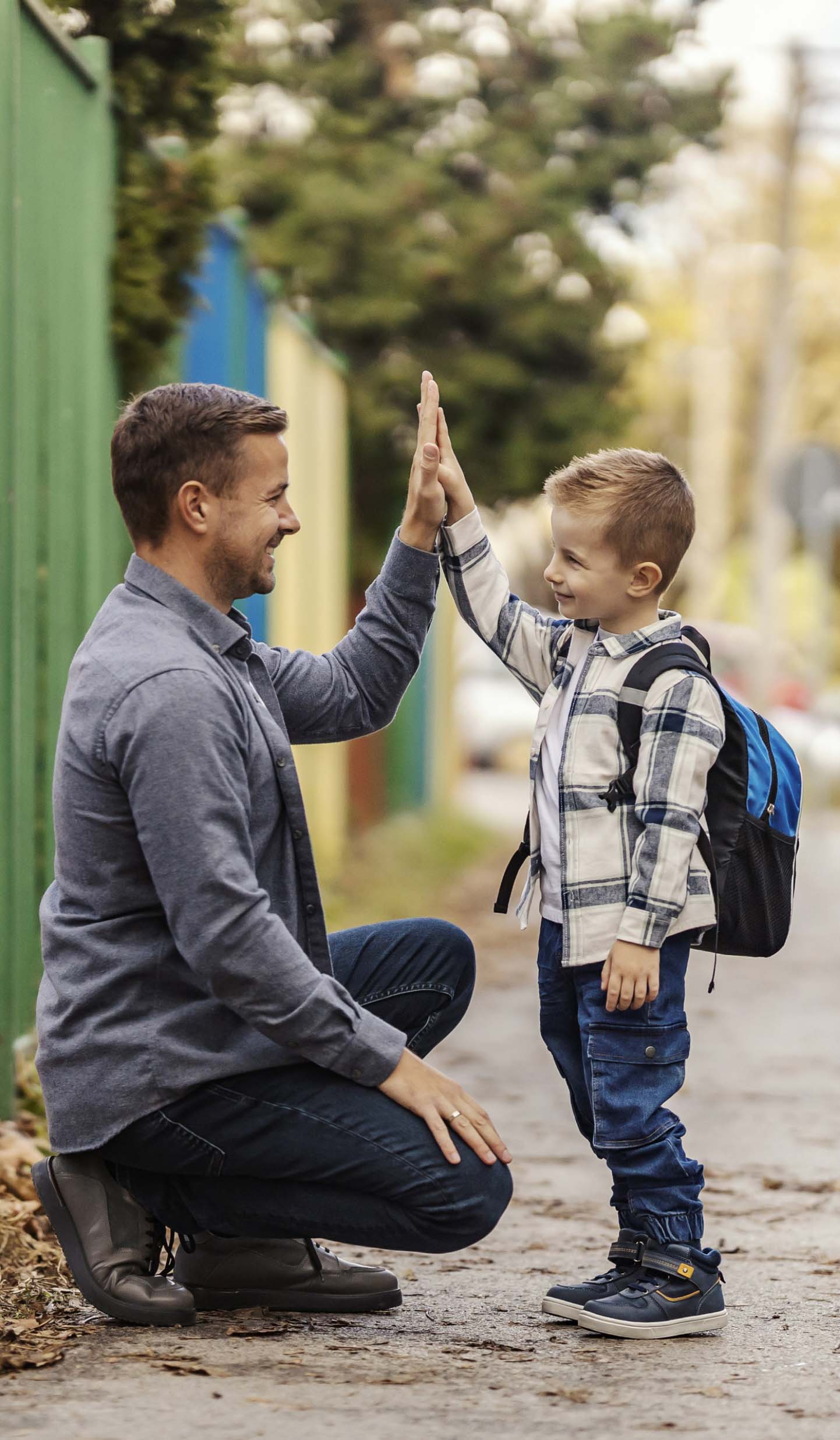 Father kneeled down high-fiving his young backpack-wearing son outside on a sidewalk