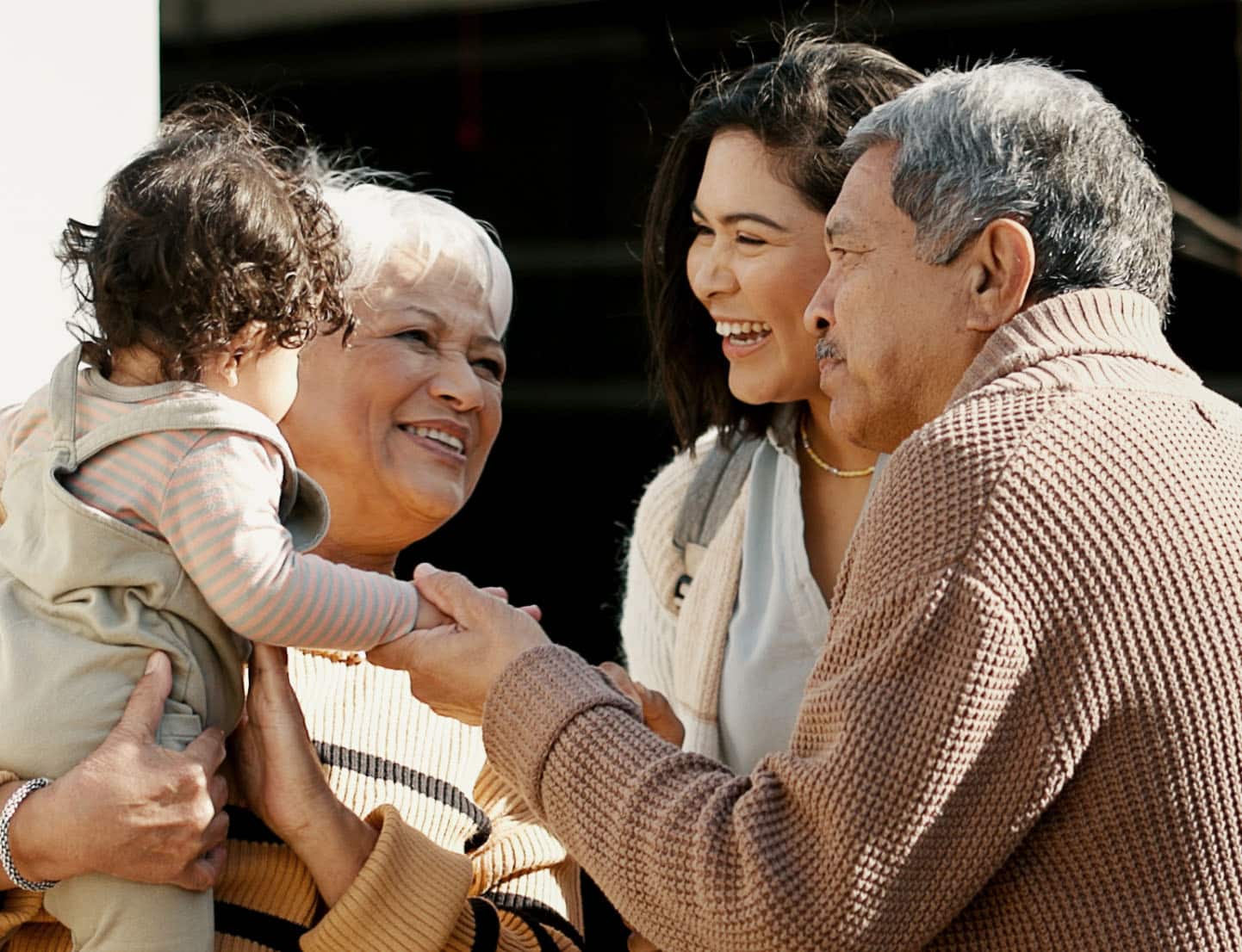 A three-generation family is smiling and interacting outdoors. A baby is held by an older woman with short white hair, while a younger woman and an older man stand beside them, looking on and holding the baby's hand.